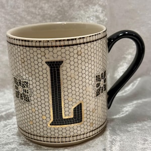 May include: A ceramic mug with a black handle and a cream-colored body. The mug features a black letter "L" with gold accents, surrounded by a mosaic-like pattern. Black floral designs and gold dots add detail.