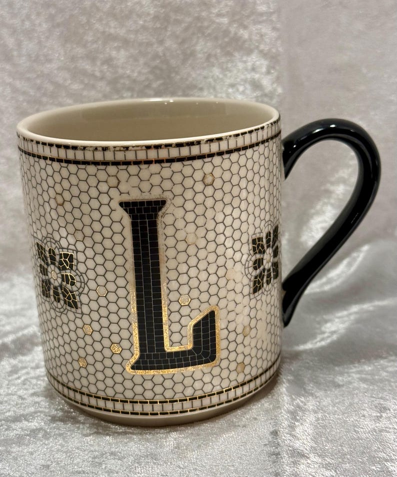 May include: A ceramic mug with a black handle and a cream-colored body. The mug features a black letter "L" with gold accents, surrounded by a mosaic-like pattern. Black floral designs and gold dots add detail.