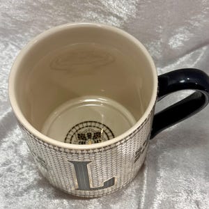 May include: Cream-colored ceramic mug with a black handle. The mug features a textured, honeycomb-like pattern with the letter "L" in a dark gray color. The interior is cream-colored, and the bottom has a decorative design.
