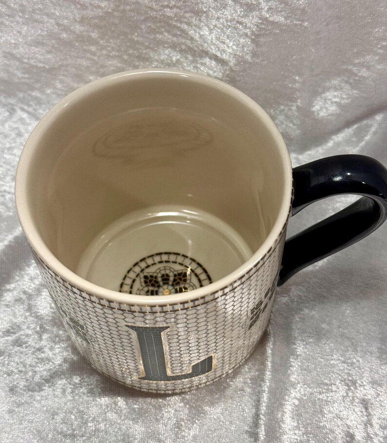 May include: Cream-colored ceramic mug with a black handle. The mug features a textured, honeycomb-like pattern with the letter "L" in a dark gray color. The interior is cream-colored, and the bottom has a decorative design.