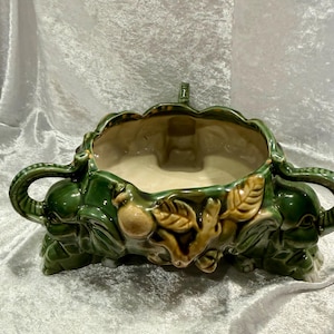May include: A green ceramic planter with elephant-shaped handles and feet. The planter features relief details of leaves and fruit. The interior is a light beige color, and the exterior is a glossy green.