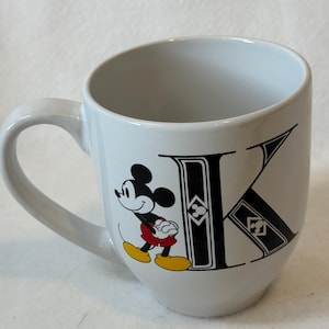May include: White ceramic mug featuring Mickey Mouse and the letter "K" in black. Mickey is depicted in his classic outfit, with red shorts and yellow shoes. The mug has a curved design and a white handle.