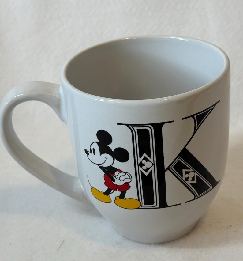 May include: White ceramic mug featuring Mickey Mouse and the letter "K" in black. Mickey is depicted in his classic outfit, with red shorts and yellow shoes. The mug has a curved design and a white handle.