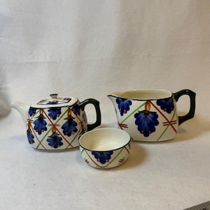 May include: A white ceramic tea set with a teapot, creamer, and small bowl. The set features a blue floral design with green and red accents. The teapot has a dark green handle and lid. The creamer and bowl also have green handles and rims.