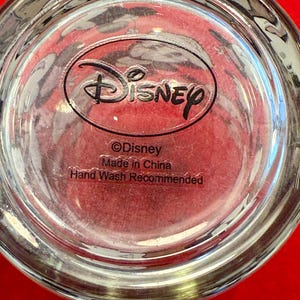 Puede incluir: Vaso transparente con el logotipo de Disney y texto. El texto incluye "&copy;Disney", "Made in China" y "Hand Wash Recommended". El vaso est&aacute; sobre una superficie roja.
