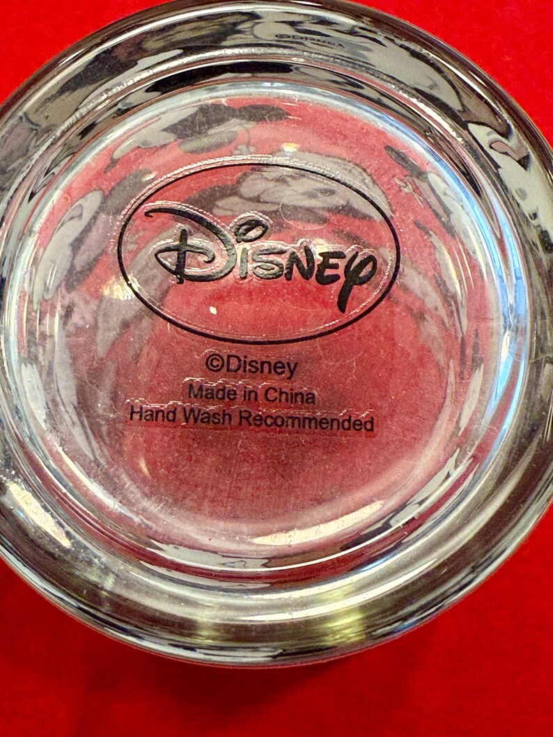 Puede incluir: Vaso transparente con el logotipo de Disney y texto. El texto incluye "&copy;Disney", "Made in China" y "Hand Wash Recommended". El vaso est&aacute; sobre una superficie roja.