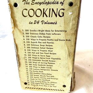 May include: Vintage cookbook titled "The Encyclopedia of Cooking in 24 Volumes." The cover is a light beige with black text and a decorative border. The book lists various recipes, including snacks, cakes, and desserts.