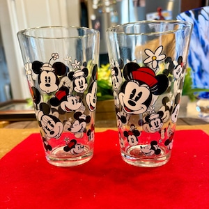 Puede incluir: Dos vasos transparentes con un patr&oacute;n repetido de caras de Mickey y Minnie Mouse, con detalles en rojo y negro. Los vasos est&aacute;n sobre una superficie roja.