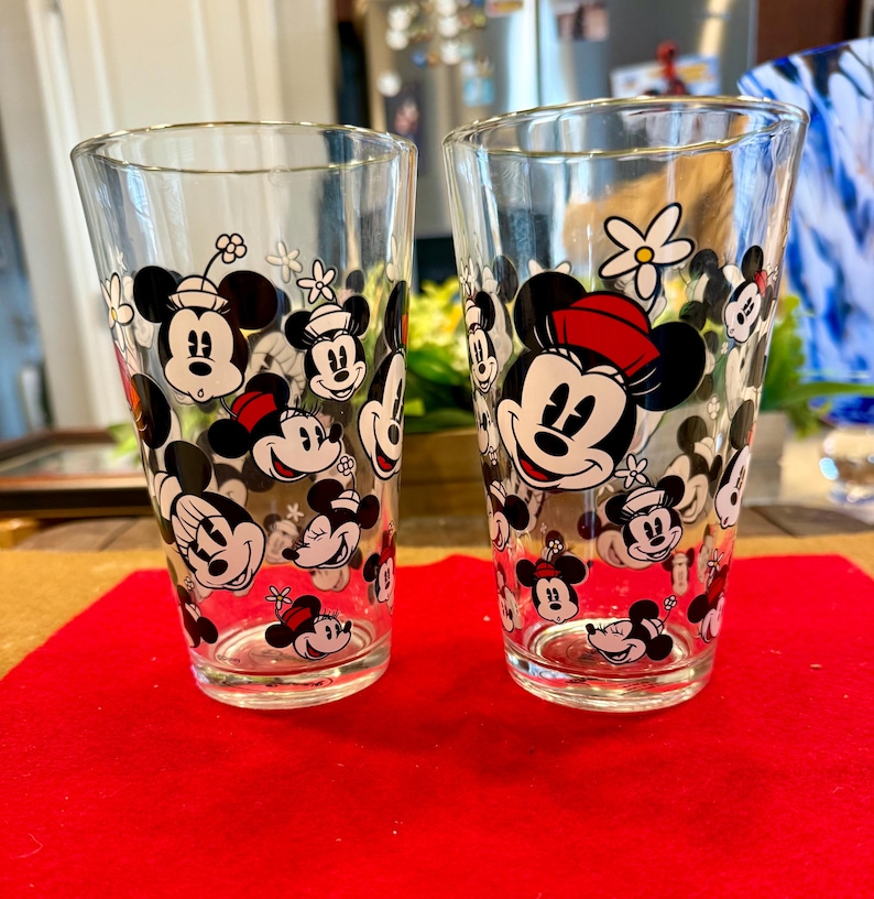 Puede incluir: Dos vasos transparentes con un patr&oacute;n repetido de caras de Mickey y Minnie Mouse, con detalles en rojo y negro. Los vasos est&aacute;n sobre una superficie roja.