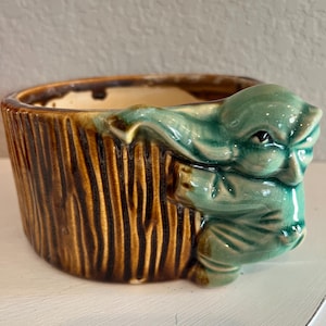 May include: A brown and green ceramic planter shaped like a tree trunk with a green gnome figure climbing up the side.