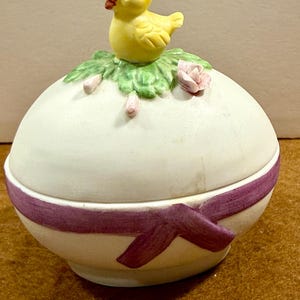 May include: A ceramic Easter egg-shaped trinket box. The lid features a yellow chick perched on green leaves with pink accents. The egg is white with a purple ribbon design around the base. A decorative item for Easter.