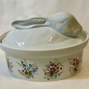 May include: A white ceramic lidded casserole dish featuring a sculpted rabbit figure on the lid. The exterior of the round base is decorated with a repeating pattern of colorful wildflowers, including red, blue, and yellow blossoms with green leaves.