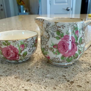 May include: A vintage cream pitcher and sugar bowl set. The white porcelain pieces feature pink rose floral designs with green leaves and gold trim. The sugar bowl is round, and the pitcher has a handle and spout.