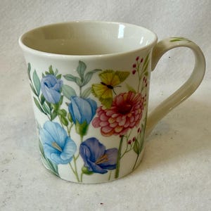 May include: Cream-colored ceramic mug with a floral design. The mug features blue, pink, and yellow flowers, along with green leaves and a yellow butterfly. The handle is cream-colored with green leaf accents. A decorative item for the kitchen.