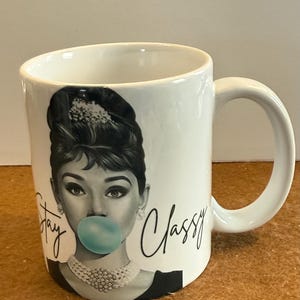 May include: White ceramic mug featuring a black and white image of a woman with a bubblegum bubble. The words "Stay Classy" are written in black script. The mug has a white handle.