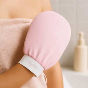 May include: A pale pink exfoliating mitt with a white wristband bearing the brand name "Glydelle". The mitt is worn on an arm, implying bathroom use. A loop is attached to the wristband for hanging.