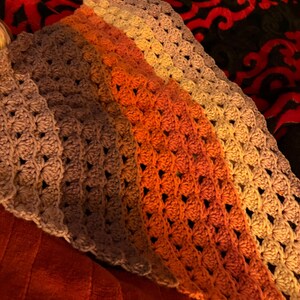 May include: A handmade crochet shawl in shades of orange, brown, and tan. The shawl features a textured stitch pattern and a triangular shape. The yarn colors transition from dark to light, creating a gradient effect. The shawl is likely made of soft yarn.