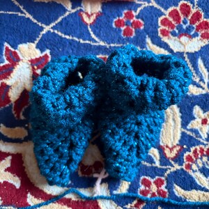 May include: A pair of teal blue crochet baby booties. The booties are made with a textured stitch and have a sparkly appearance. A pink crocheted headband is visible in the background. The booties are on a patterned rug.