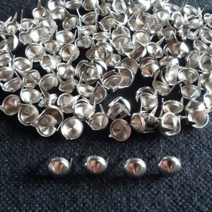 May include: A collection of small, shiny silver upholstery tacks with a rounded head and a pointed tip. The tacks are scattered across a dark, textured surface, with a few arranged in a row.
