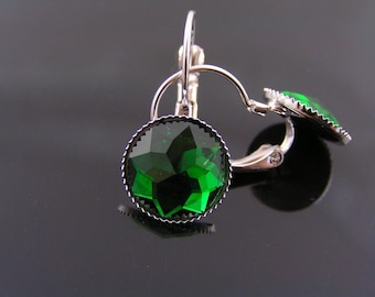 May include: A pair of leverback earrings with large, faceted, emerald-green gemstones. The gemstones are set in a silver-colored metal with a decorative, scalloped edge. The earrings are displayed against a dark, reflective surface.