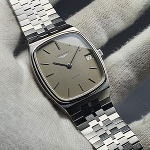 Pode incluir: Um relógio Longines Quartz prateado com mostrador retangular e pulseira de elos prateados. O mostrador do relógio é de cor champanhe clara com ponteiros e marcadores de hora pretos. A data é exibida às 3 horas.