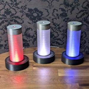 May include: Three cylindrical LED lamps with silver tops and black bases. The lamps are lit with red, white, and blue light, respectively. The lamps are on a wooden surface.