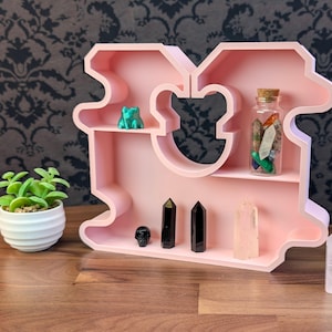 May include: A pink, bear-shaped shelf with multiple shelves, holding a small green frog figurine, a glass jar filled with crystals, and various crystals and decorative objects. A small potted plant sits to the left.
