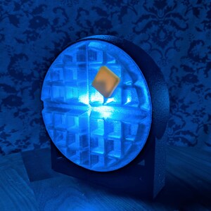 May include: A unique, illuminated waffle-shaped lamp. The lamp emits a bright blue light, with a square, yellow-orange accent in the center. The lamp is set on a black base, creating a striking contrast against the blue light.