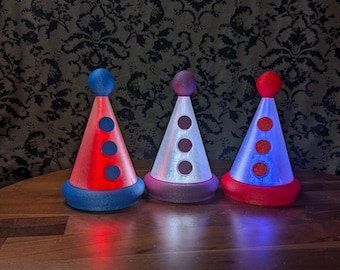May include: Three cone-shaped decorative objects with rounded tops and circular accents. The left object is red with a blue base and top. The center object is white with a purple base and top. The right object is blue with a red base and top.