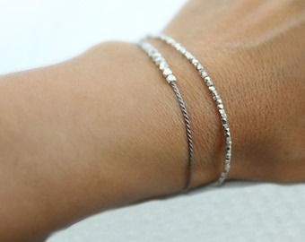 Könnte beinhalten: Zwei silberne Armbänder am Handgelenk einer Person. Ein Armband ist eine dünne Kette mit kleinen silbernen Perlen. Das andere Armband ist eine dickere Kette mit größeren silbernen Perlen.