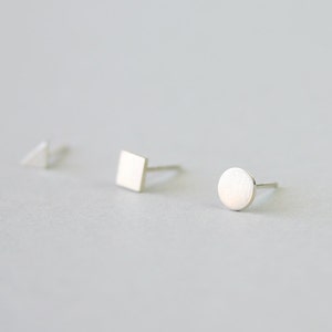 Può includere: Tre orecchini a lobo in argento di forma geometrica: un triangolo, un quadrato e un cerchio. Gli orecchini sono piccoli e minimalisti, con finitura in metallo spazzolato. Sono esposti su una superficie grigio chiaro.