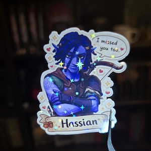 May include: A holographic sticker featuring a blue-skinned character with blue hair and pointed ears. The character is holding a bow and arrow. Text on the sticker reads "Hassian" and "I missed you too."