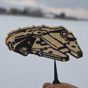 Può includere: Un adesivo beige e nero della navicella spaziale Millennium Falcon con dettagli di stelle olografiche. L'adesivo è tenuto da uno strumento nero su uno sfondo di cielo nuvoloso e neve.