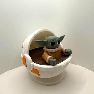 May include: A figurine of a character from a popular science fiction series. The figure has large ears, a tan coat, and is seated in a white and orange cradle. The cradle has a brown interior and is on a white base.