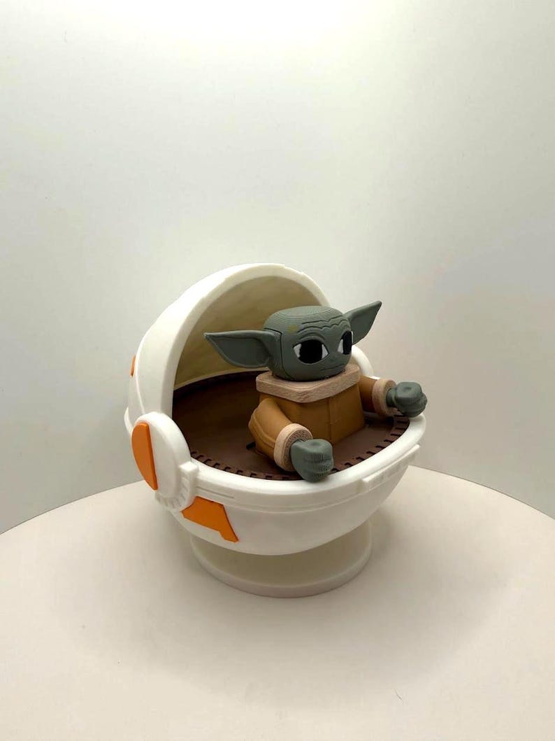 May include: A figurine of a character from a popular science fiction series. The figure has large ears, a tan coat, and is seated in a white and orange cradle. The cradle has a brown interior and is on a white base.