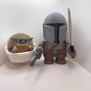 May include: A pair of Star Wars-themed figurines. One is a Mandalorian figure in gray and brown armor, holding a blaster and a staff. The other is a Grogu figure in a white and orange cradle. The figures are on a white surface.