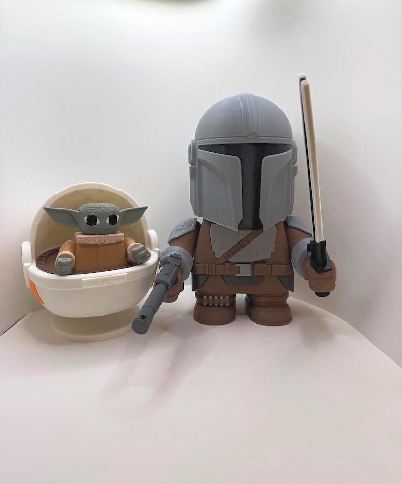 May include: A pair of Star Wars-themed figurines. One is a Mandalorian figure in gray and brown armor, holding a blaster and a staff. The other is a Grogu figure in a white and orange cradle. The figures are on a white surface.
