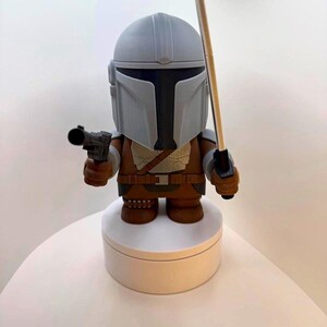 May include: A Mandalorian figure with a gray helmet and brown armor, holding a blaster pistol and a staff. The figure is standing on a white circular base. The figure is a stylized representation of the character.