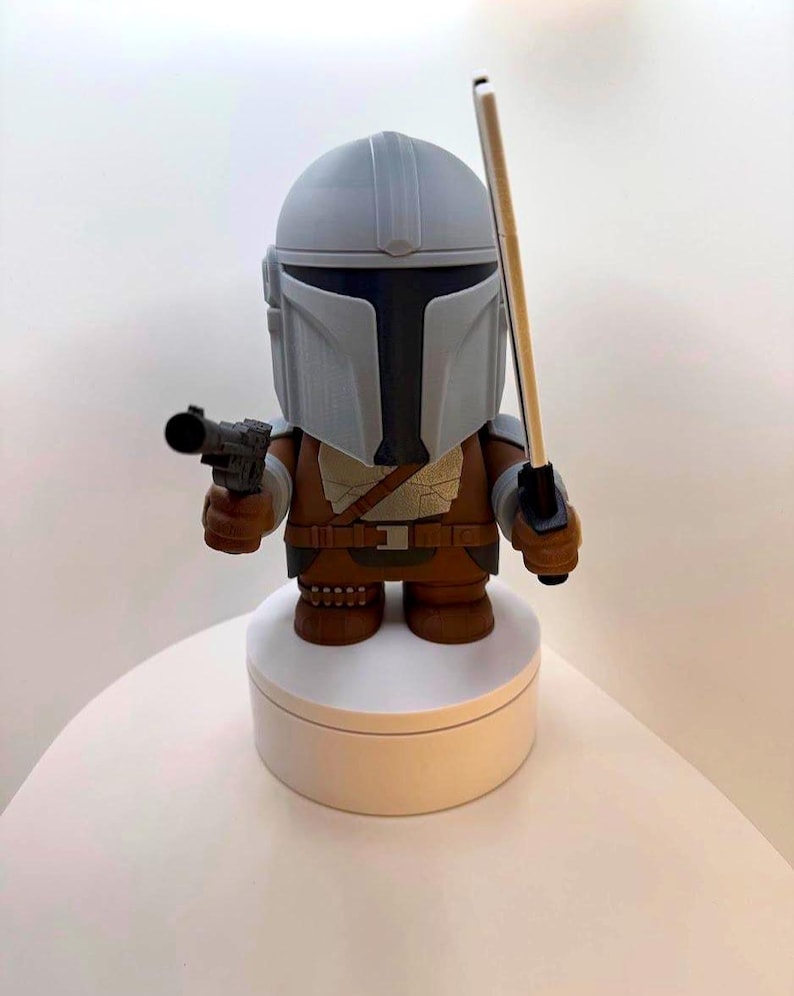 May include: A Mandalorian figure with a gray helmet and brown armor, holding a blaster pistol and a staff. The figure is standing on a white circular base. The figure is a stylized representation of the character.