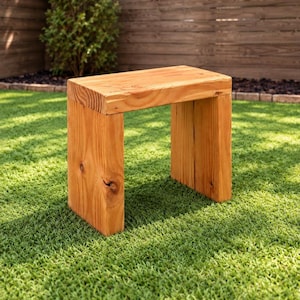 May include: A rectangular wooden stool constructed from thick, light brown planks with visible wood grain and natural knots. The stool features a flat top surface supported by two vertical side panels, creating a simple, open-frame structure.