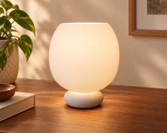 May include: A spherical table lamp with a frosted white glass shade and a smooth, rounded white base. The lamp emits a soft, warm glow, illuminating the surface it rests upon. The design features a simple, clean silhouette with horizontal texture.