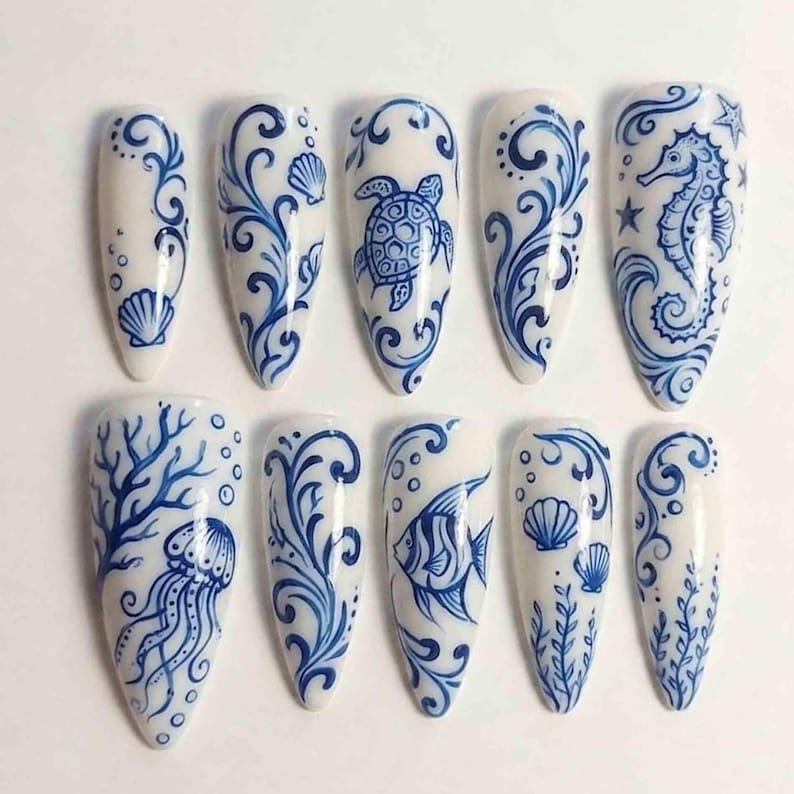 Puede incluir: Un conjunto de diez u&ntilde;as postizas blancas en forma de almendra con intrincados dise&ntilde;os azules. Cada u&ntilde;a presenta una ilustraci&oacute;n diferente con tem&aacute;tica marina, incluyendo un caballito de mar, una tortuga, un pez, una medusa y conchas, con remolinos y patrones decorativos.