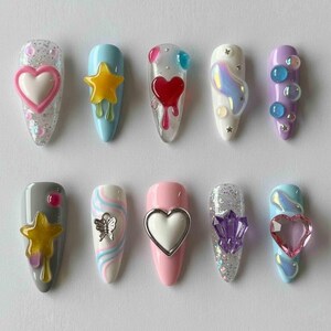 May include: A collection of ten artificial nails with various designs. The nails feature a range of colors, including pink, blue, and gray, and are adorned with heart, star, and butterfly embellishments. Some nails have glitter accents and 3D elements.