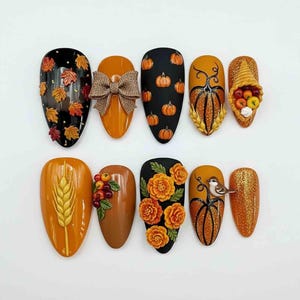Può includere: Una collezione di dieci unghie finte a forma di mandorla, decorate. I disegni includono temi autunnali come zucche, foglie, grano, fiori e una cornucopia. I colori sono nero, arancione, marrone e oro, con abbellimenti 3D.