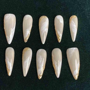 May include: Ten almond-shaped artificial nails. Each nail is a creamy beige color with white marbling and a gold foil accent along one side. The nails are arranged on a dark green surface.