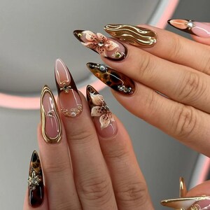 May include: A set of long, almond-shaped acrylic nails with intricate designs. Some nails feature 3D floral art, others have gold accents, and some showcase a tortoiseshell pattern. The color palette includes nude, brown, and gold.