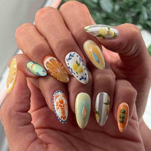 May include: A hand with a manicure featuring various food-themed designs. The nails showcase detailed illustrations of lemons, oranges, fish, and a lobster, with a mix of colors including orange, yellow, blue, and green. The nail shapes are oval.