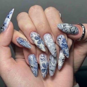 May include: Almond-shaped nails featuring a blue and white color palette with intricate floral and swirling patterns. The nail art includes hand-painted designs and small, clear, circular rhinestones applied to several of the nail surfaces.