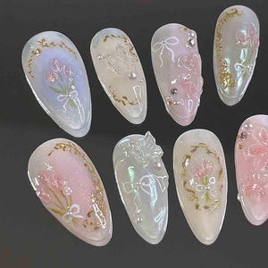 Op de afbeelding: Een verzameling kunstnagels met diverse ontwerpen. Sommige nagels tonen roze en witte bloemstukken met gouden accenten. Andere hebben strikken, parels en heldere, getextureerde versieringen. De nagels zijn ovaal en hebben een glanzende afwerking.