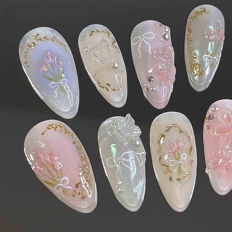 Op de afbeelding: Een verzameling kunstnagels met diverse ontwerpen. Sommige nagels tonen roze en witte bloemstukken met gouden accenten. Andere hebben strikken, parels en heldere, getextureerde versieringen. De nagels zijn ovaal en hebben een glanzende afwerking.
